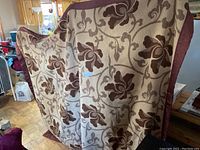 Photo showing a large wool blanket with a cream background and brown floral and vine pattern, bordered in brown fabric.