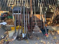 Photo showing an assortment of yard tools including shovels, rakes, and cutters lined up against a wooden backdrop with dry leaves on the ground