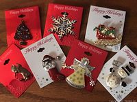 Photo shows 7 holiday-themed pins arranged on red and white 'Happy Holidays' backing cards. Pins include a Christmas tree, snowflake, Santa in stocking, sleigh with gifts, reindeer, angel, and snowman.