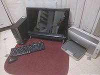 Photo of computer setup showing desktop tower, widescreen LCD monitor, wired keyboard and mouse, and HP inkjet printer on maroon surface.