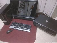 Photo of a black HP desktop tower, HP monitor, wired keyboard, wired mouse and HP all-in-one printer on a red surface.