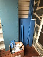 Blue metal shelving panels and vertical supports leaning against wall with box of assembled hardware.