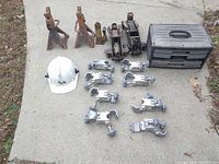 Photo showing two rusty orange metal jack stands, two black hydraulic jacks, eight metal special clamps with multiple adjustable knobs, a white hard hat with TDA logo, and a black plastic toolbox.
