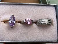 Three rings displayed in a soft, light pink cushioned jewelry box, showing different angles and details of each ring's gemstone and band.