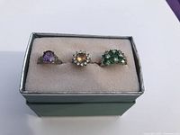 Three rings displayed in a green box with beige cushion. Front view shows amethyst, citrine surrounded by clear stones, and green stones rings.