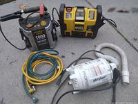 Photo showing two Stanley jump boxes, a white Stryker Propelair blower, and four hoses (green, yellow, white). The jump boxes have attached jumper cables. Items are placed on a concrete surface.