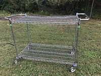 Full view of large metal rolling wire cart with two shelves and handles on each end, standing on grass outdoors.