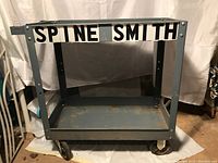 Front and side views of a gray metal utility cart with two shelves and four caster wheels showing rust and wear on shelves.