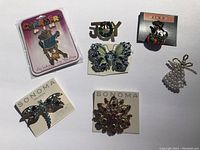 Photo showing a collection of 7 assorted decorative pins on a white surface, including a Halloween cat in pumpkin pin, butterfly pins, dragonfly, flower, an angel, and a JOY lettering pin.