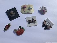 Image showing seven decorative pins arranged on solid background, featuring a motorcycle, flamingo, pumpkin, butterfly, flower pins, and a silver-tone teapot pin.