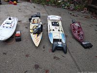 Four R/C boats in a row on a concrete surface, showing different models and two remote controllers, plus a battery charger on the side.