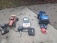 Photo showing the three R/C cars (red dune buggy, white car, blue monster truck) with their respective controllers and a red helmet accessory arranged on a concrete surface outdoors.