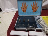 Open case showing CT.ion therapy device with three chrome knobs, connection ports, probes, and hand electrode straps. Diagram of hand acupoints visible on lid.