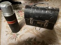 Black metal antique lunch box with handle and matching thermos placed on carpeted surface