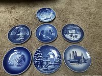 Seven 1970s blue and white collector porcelain plates laid out on carpet. Five feature Christmas and winter scenes, two German themed, including Olympic commemorative.