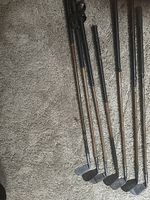 Seven antique golf clubs with hickory shafts and dark grips, seen from above resting on carpet.