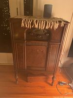 Front view of antique wooden console radio cabinet showing front speaker grille with fabric and radio controls above.
