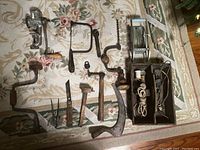 Wide shot of assortment of antique hand tools arranged on a floral rug including braces, saws, hammers, pliers, and wooden box with electrical parts.