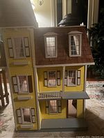 Front exterior view of three-story yellow wooden doll house with brown shingled roof and multiple windows.