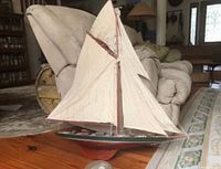 Full view of the large reproduction sail boat model showing cloth sails, mast rigging, wood deck, and painted hull on a wood floor next to furniture.