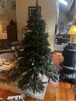 Front view indoor photo of a 7.5 foot tall green artificial Christmas tree set up in a living room with wooden flooring and furniture surrounding. Tree is upright, unlit, needle foliage visible, three-piece assembly confirmed.
