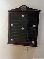 Front view of the wooden cherry golf ball display cabinet mounted on a white wall, showing some golf balls in a few slots but mostly empty.