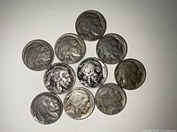 Top-down view of 10 Buffalo nickels arranged in a circular pattern with readable dates, showing various levels of wear and tarnish on each coin.