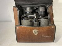 Open case revealing black textured Bushnell binoculars with attached straps inside the brown leather case with Bushnell logo.