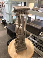 Full view of the tall decorative pedestal showing the candle platform, column with fluted design, and cherub figures at the base. The pedestal has a tassel rope accessory.