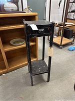 Front angled view of a black painted wooden plant stand with a drawer and a lower shelf, showing wear and chipping paint.
