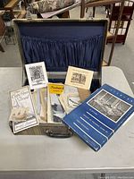 Open suitcase showing assorted paper ephemera including brochures, booklets, and prints on a table.