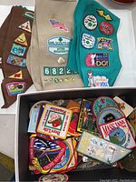 Three fabric sashes laid out with sewn-on badges and pins, a beige sash with an American flag patch and troop numbers, a brown sash with multiple triangular badges, and a teal sash with colorful badges embroidered with phrases and animals.
