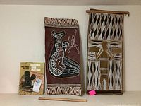 Wide shot of both bark paintings and the book together on a white shelf, showing the relative size and condition of all three items.