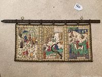Full view of the quilted wall hanging showing three embroidered panels with Arabic scenes, bordered by decorative edging, hanging on the carved wooden mounting rod with rings.