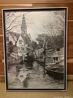 Full view of framed ink on paper art print showing river, houses, and church steeple.