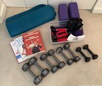 Photo showing a rolled blue yoga mat, two purple foam yoga blocks, a clear plastic container with red and purple dumbbells and exercise bands, and exercise books including Arnold Schwarzenegger's 'Bodyshaping For Women'