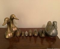 Single image showing full set of 9 metal ducks including the pair of brass bookends on the left, various smaller brass and metal duck figurines arranged in a line, and the large painted duck trinket box on the right.