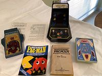 Wide view showing all 5 items: Pac-Man handheld arcade game, Wildfire handheld game with instructions, Rocket Pinball handheld game, and two vintage Pac-Man strategy books.