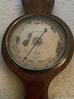 Close-up of enamel barometer dial showing scale markings, pointer, brand inscriptions and surface patina.