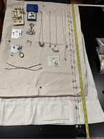 Top-down photo showing multiple silver-toned necklaces with various pendants, earrings, bracelets, pin, and a key fob arranged on cloth.