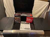 Photo showing the open grill with side shelves extended, grill grates, and multiple instructional and promotional booklets for Tru-Infrared technology placed inside.