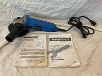 Angle grinder laid flat showing blue body, black side handle, power cord, grinding disc, metal removal tool, and two manuals (owner's and instruction).