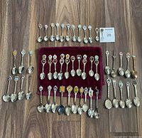 Overview of the 51 collector spoons arranged in rows on a wooden floor with one mini fork included, showing a mix of enamel and metal designs.