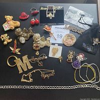 Full lot showing assorted costume jewelry including pins shaped like teddy bear, red hat, cherries, Christmas stocking, angels, and floral designs. Includes necklaces with name pendants, keychain with 'M', earrings, and adjustable ring.