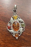 Front view of silver pendant featuring central large oval honey amber stone surrounded by triangular stones in lemon, green, red, and honey amber hues and intricate leaf silver detailing.