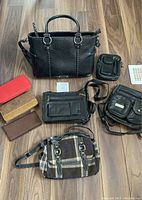 Overview of assorted purses and wallets on floor showing variety of styles, colors, and sizes including black leather, plaid, and brown wallets.