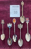 Six collectible spoons displayed on a burgundy fabric background, showing distinct decorative tops and handles with themed designs.