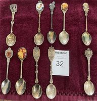 Full lot of 10 commemorative royal family collector spoons displayed on red velvet background, showing different designs including portraits, crests, and inscriptions.