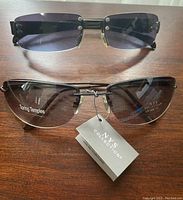 Two pairs of sunglasses on a wooden surface: one new NYS Collection pair with tag and one used pair with dark rectangular lenses and teal arms