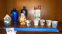 Full lot view showing figurines, vases, sake flask, and five cups on wooden shelf.